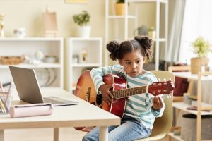 Black little girl playing guitar at home and watching online music lesson