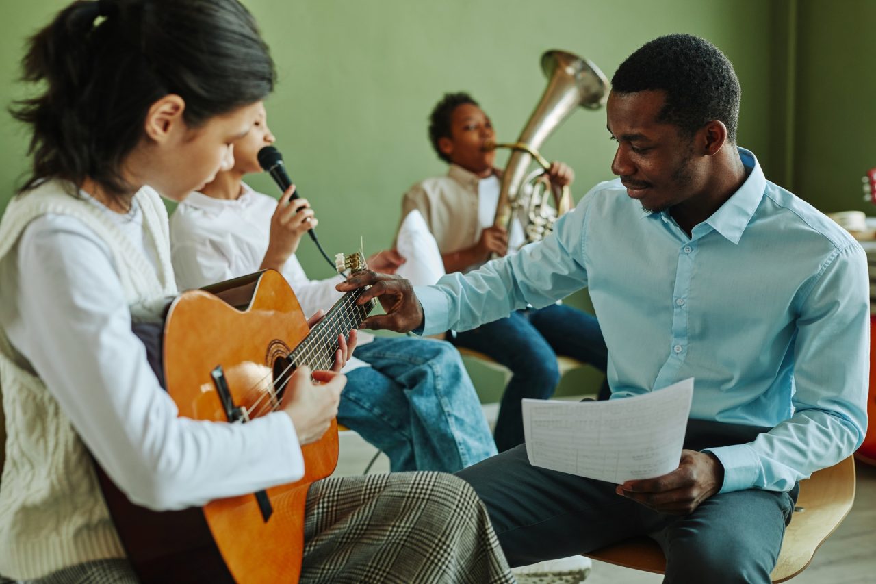 Young teacher of music consulting Asian schoolgirl with acoustic guitar